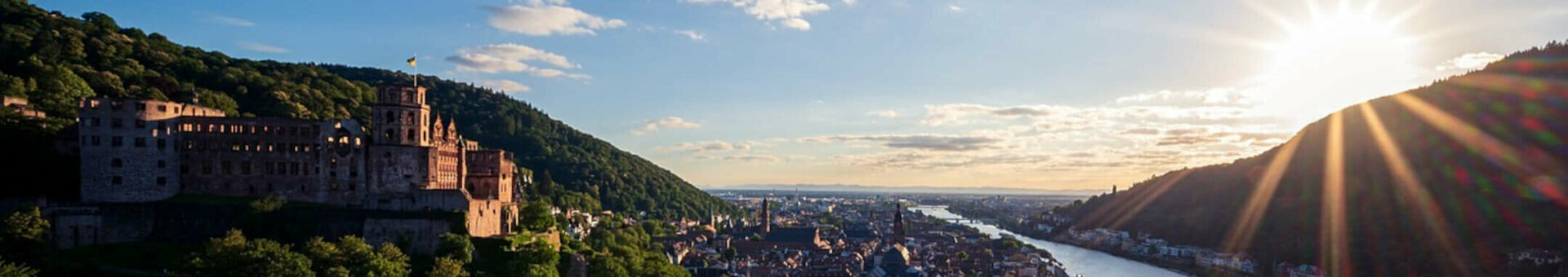 View of the castle and town by the river at sunset, surrounded by green hills.