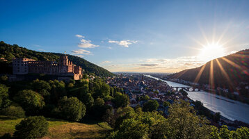 Blick auf Schloss und Stadt am Fluss bei Sonnenuntergang, umgeben von grünen Hügeln.