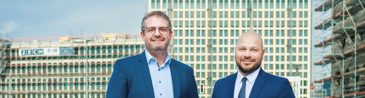 ATLANTIC Hotel Heidelberg | Hoteldirektor Stephan Sporer und Verkaufsleiter Stefan Knittel Zwei Männer in Anzügen stehen lächelnd vor einem im Bau befindlichen Gebäude.