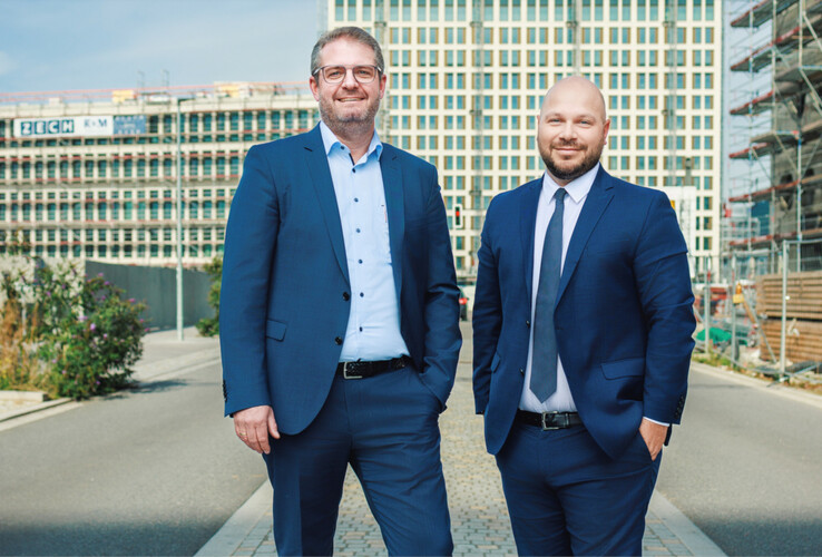 Zwei Männer in Anzügen stehen lächelnd vor einem im Bau befindlichen Gebäude.