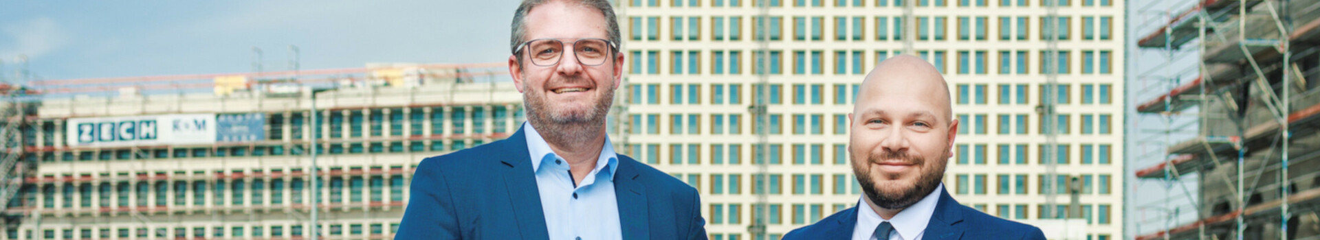 Zwei Männer in Anzügen stehen lächelnd vor einem im Bau befindlichen Gebäude.