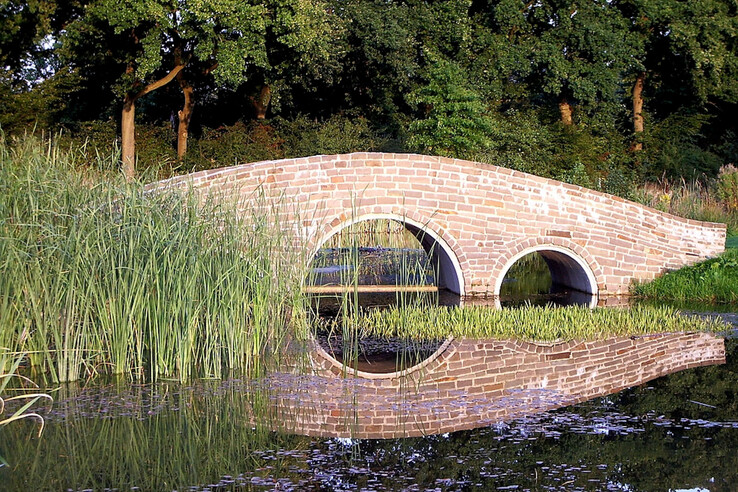 Steinbrücke mit zwei Bögen über einem Teich, umgeben von Schilf und Bäumen.