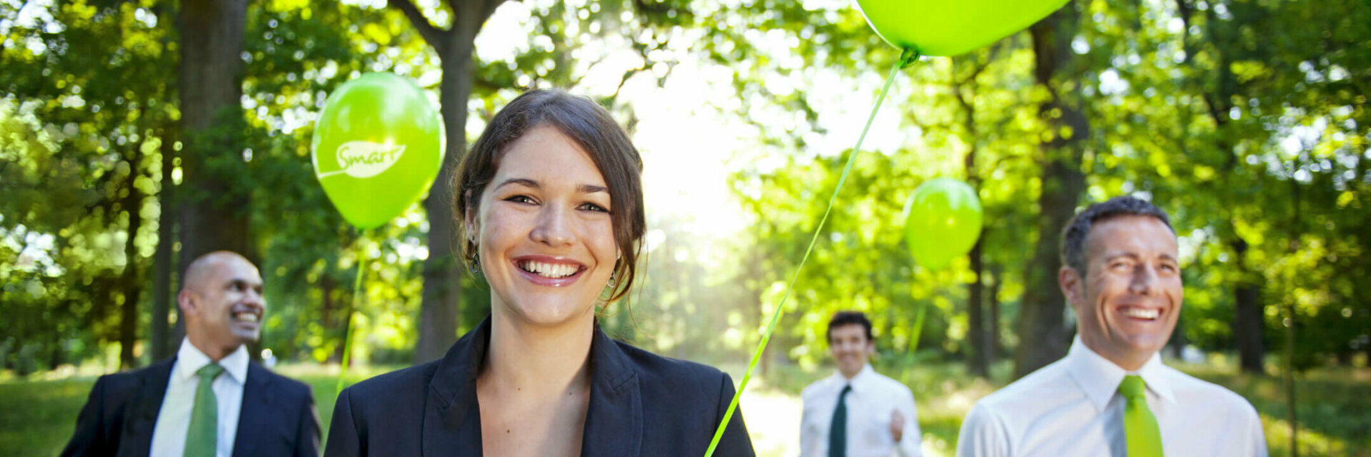 Lächelnde Menschen in Anzügen halten grüne Ballons im sonnigen Wald.