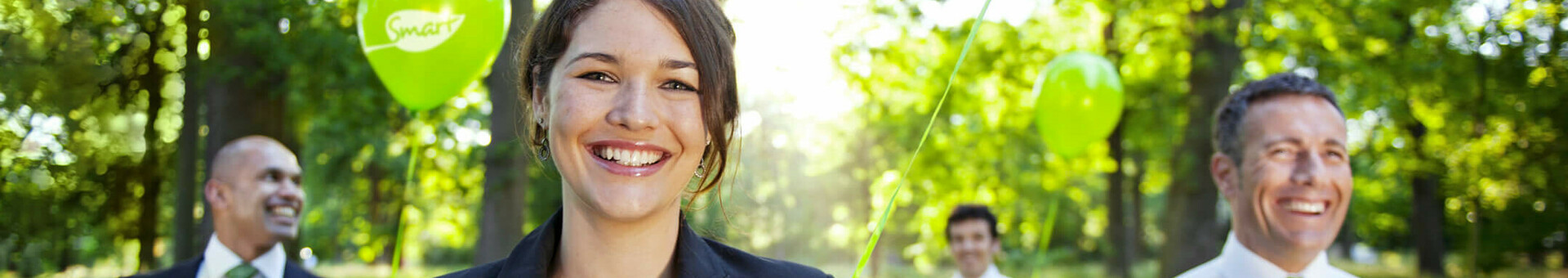 Smiling people in suits hold green balloons in the sunny forest.