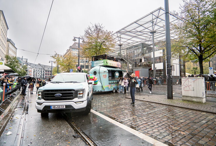 Straßenszene mit weißem Truck, Anhänger, Menschenmengen und regnerischem Wetter in einer belebten Stadtumgebung.