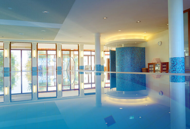 Helles Hallenbad mit großen Fenstern, blauem Mosaik, Liegestühlen und frischen Handtüchern auf einem Regal.