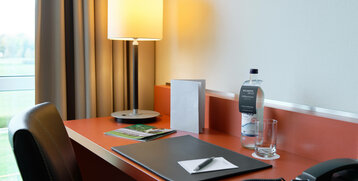 Desk in ATLANTIC hotel room with lamp, water bottle, glass, notepad and telephone, window view of green space.
