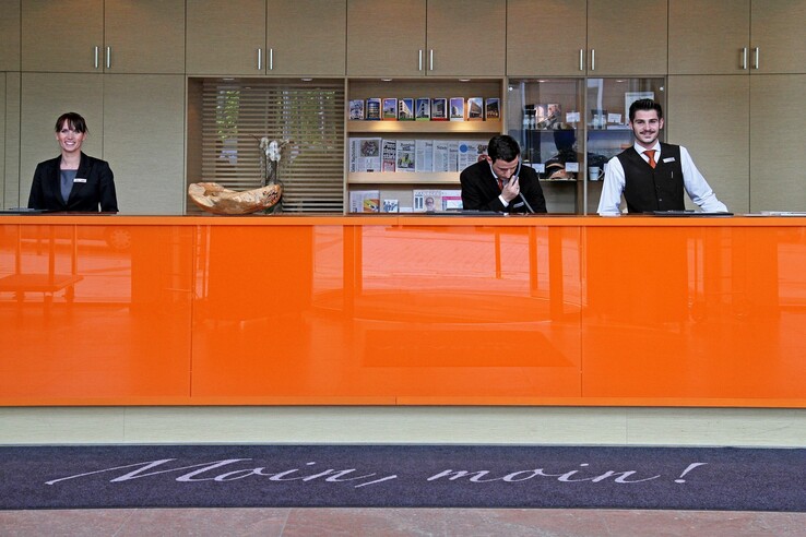 Hotelrezeption mit drei Mitarbeitenden hinter einem orangefarbenen Tresen, begrüßender Schriftzug "Moin, moin!" auf Teppich.