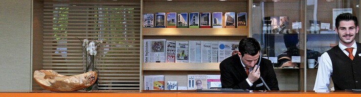 Wir bieten Ihnen erstklassigen Service und First Class-Komfort zu fairen Preisen Hotelrezeption mit drei Mitarbeitenden hinter einem orangefarbenen Tresen, begrüßender Schriftzug "Moin, moin!" auf Teppich.