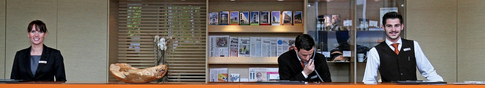 Hotelrezeption mit drei Mitarbeitenden hinter einem orangefarbenen Tresen, begrüßender Schriftzug "Moin, moin!" auf Teppich.