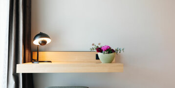 Minimalist hotel room with wooden table, table lamp and flower vase, next to a gray stool.