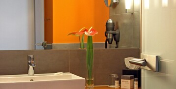 Modernes Hotelbad mit orangefarbener Wand, Waschbecken, Spiegel, Fön und frischen Blumen in Vase.