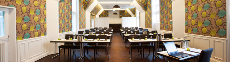 Parlimentary seating in the Scotland Saal of the ATLANTIC Grand Hotel Bremen Modern conference room in the ATLANTIC Grand Hotel Bremen with stylish wallpaper and rows of tables.