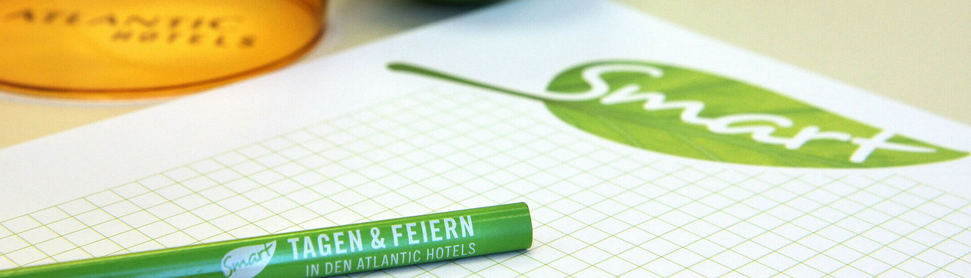 Grüner Stift und Ball mit "Smart"-Logo, orange Glas mit "Atlantic Hotels"-Schriftzug, auf kariertem Papier.