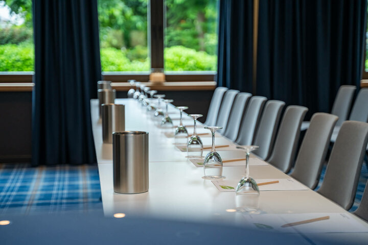 Moderner Konferenzraum mit Tisch, Stühlen, umgedrehten Gläsern und Schreibmaterial, mit Blick auf grüne Gartenanlage.