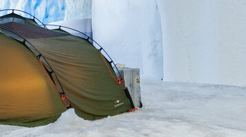 Green tent on snow in front of an icy cave wall, illuminated by warm light inside.