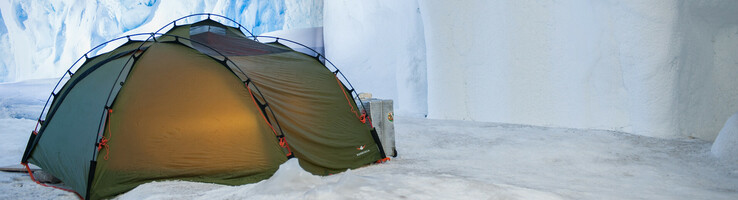 Grünes Zelt auf Schnee vor eisiger Höhlenwand, beleuchtet von warmem Licht im Inneren.