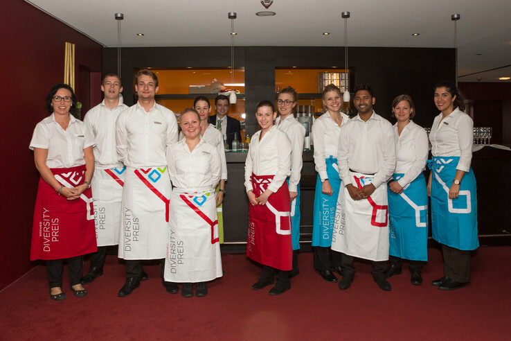 Hotel staff in white shirts and colorful aprons stand smiling in front of a bar.