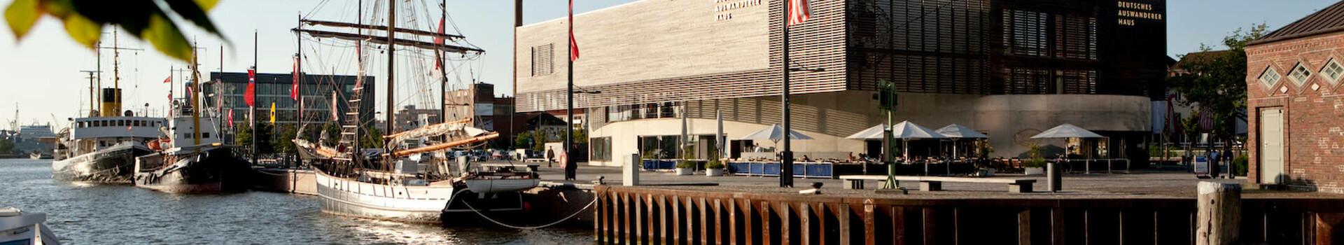 Angebot ATLANTIC Hotel Sail City | Deutsches Auswandererhaus Hafenansicht in Bremerhaven mit Segelschiffen und dem Deutschen Auswandererhaus bei Sonnenuntergang.