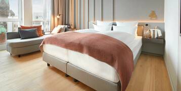 Modernes Hotelzimmer mit Doppelbett, Sofa, Holzboden und Blick auf Bremer Stadtlandschaft durch große Fenster.