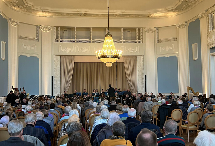 Ein Konzertsaal im ATLANTIC Grand Hotel Travemünde mit Orchester und Publikum unter einem großen Kronleuchter.