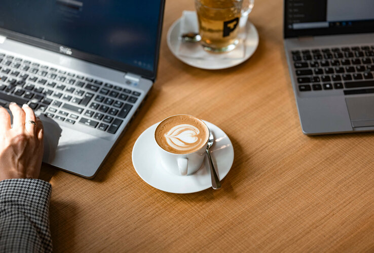 Two laptops on a wooden table, cappuccino with latte art and a glass of tea in between.