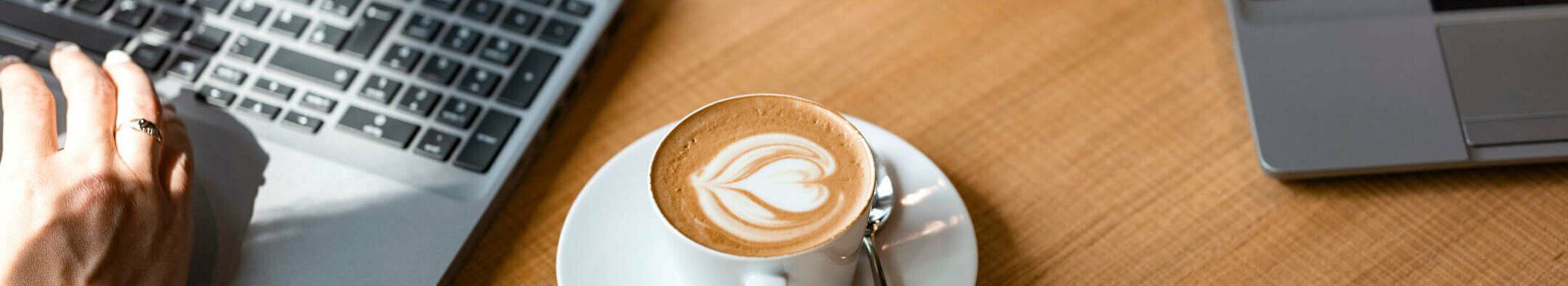 Two laptops on a wooden table, cappuccino with latte art and a glass of tea in between.