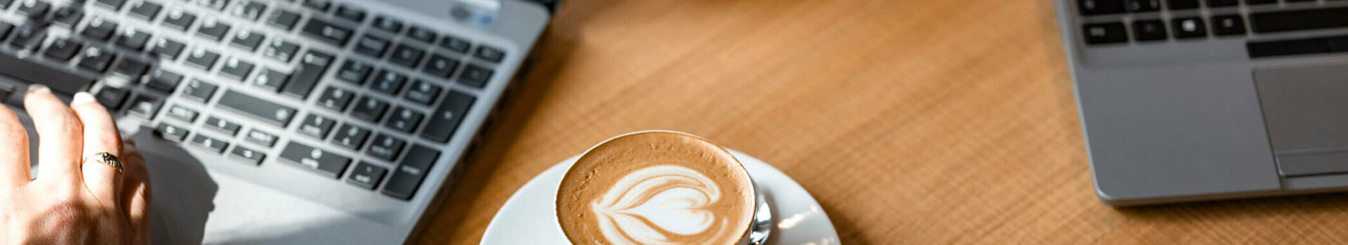 Co-Working Plätze im ATLANTIC Hotel Münster Zwei Laptops auf Holztisch, dazwischen Cappuccino mit Latte Art und Glas Tee.