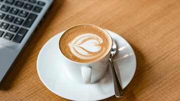 Zwei Laptops auf Holztisch, dazwischen Cappuccino mit Latte Art und Glas Tee.