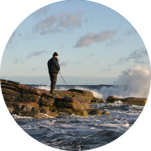 Ein Angler steht auf Felsen am Meer, während Wellen gegen die Küste schlagen.