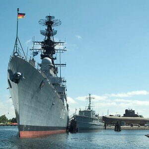 Kriegsschiff und U-Boot im Hafen, blauer Himmel, ruhiges Wasser.