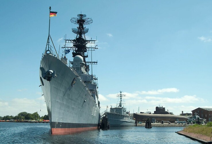 Kriegsschiff und U-Boot im Hafen, blauer Himmel, ruhiges Wasser.
