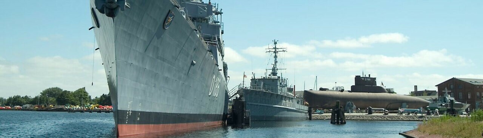 Kriegsschiff und U-Boot im Hafen, blauer Himmel, ruhiges Wasser.