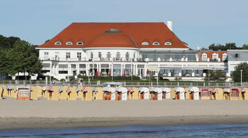 ATLANTIC Grand Hotel Travemünde mit rotem Dach, Strandkörben am Sandstrand und Meer im Vordergrund.