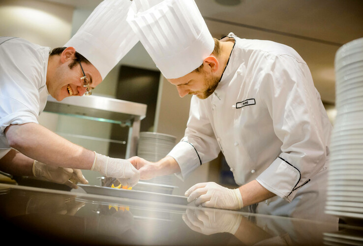 Zwei Köche in weißen Uniformen und Kochmützen richten konzentriert ein Gericht in einer Hotelküche an.