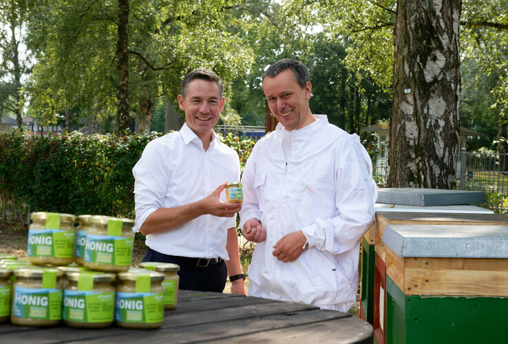 Zwei Männer lächeln, halten ein Honigglas, umgeben von Bienenstöcken und Bäumen im Freien.