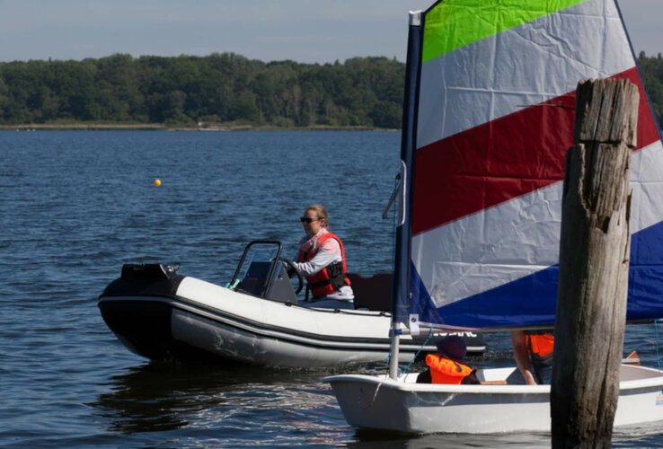 Person in motorboat and sailboat with colorful sail on a lake surrounded by forest.