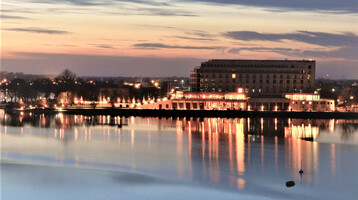 ATLANTIC Hotel Wilhelmshaven bei Dämmerung, beleuchtet, spiegelt sich im ruhigen Wasser des Hafens wider.