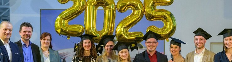 Eine Gruppe von Menschen in formeller Kleidung steht lächelnd unter goldenen "2025"-Ballons.