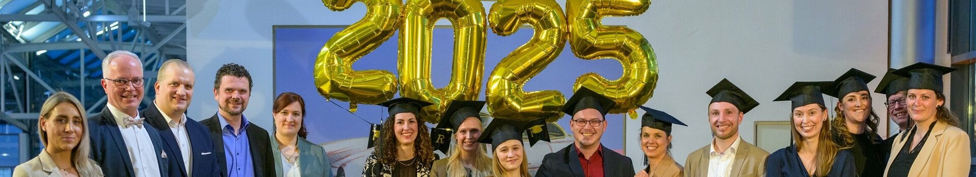 Eine Gruppe von Menschen in formeller Kleidung steht lächelnd unter goldenen "2025"-Ballons.