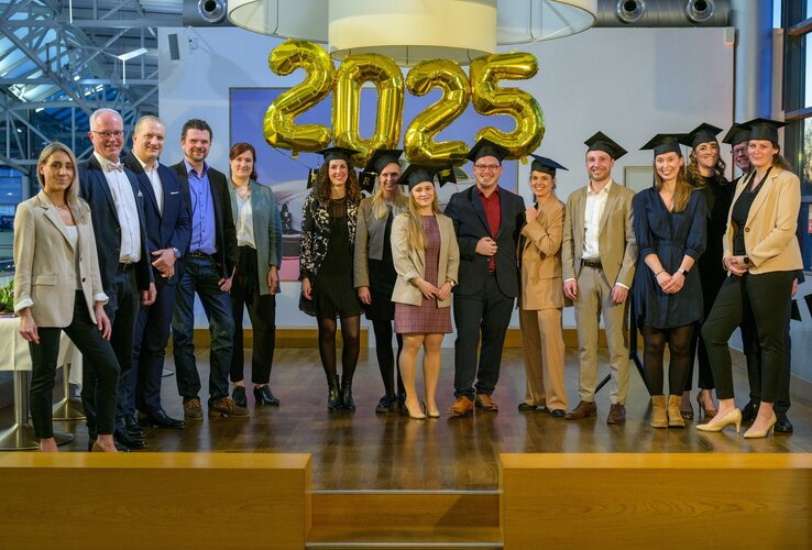 Eine Gruppe von Menschen in formeller Kleidung steht lächelnd unter goldenen "2025"-Ballons.