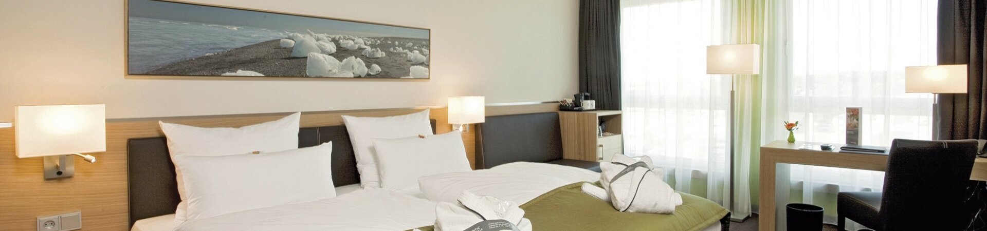 Innenansicht des Comfort Zimmer mit Doppelbett im ATLANTIC Hotel Kiel Modernes Hotelzimmer mit Doppelbett, grüner Überdecke, zwei Bademänteln, Schreibtisch und großen Fenstern.