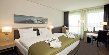 Modernes Hotelzimmer mit Doppelbett, grüner Überdecke, zwei Bademänteln, Schreibtisch und großen Fenstern.