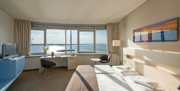 Bright hotel room with large window, view of the water, double bed, desk, armchair and modern furnishings.