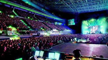 Large, illuminated concert hall with stage, audience and technical area in the foreground.
