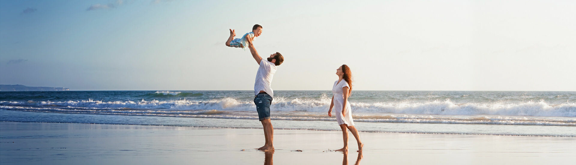 ATLANTIC Hotel Wilhelmshaven Familie am Strand; Vater hebt Kind hoch, Mutter schaut zu, im Hintergrund das Meer.