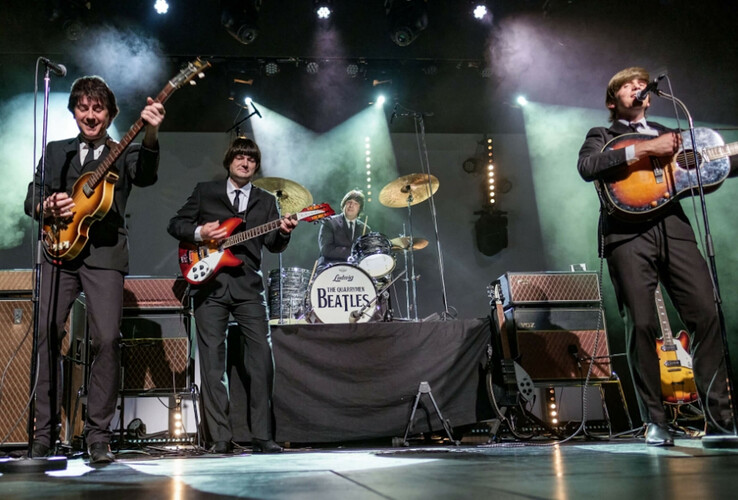 Eine Band in Anzügen spielt live auf der Bühne, umgeben von Scheinwerfern und Nebel.