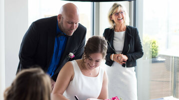Hochzeit feiern im ATLANTIC Hotel Kiel Brautpaar unterzeichnet Dokumente, während eine lächelnde Frau im Hintergrund steht, im ATLANTIC Hotel Kiel.