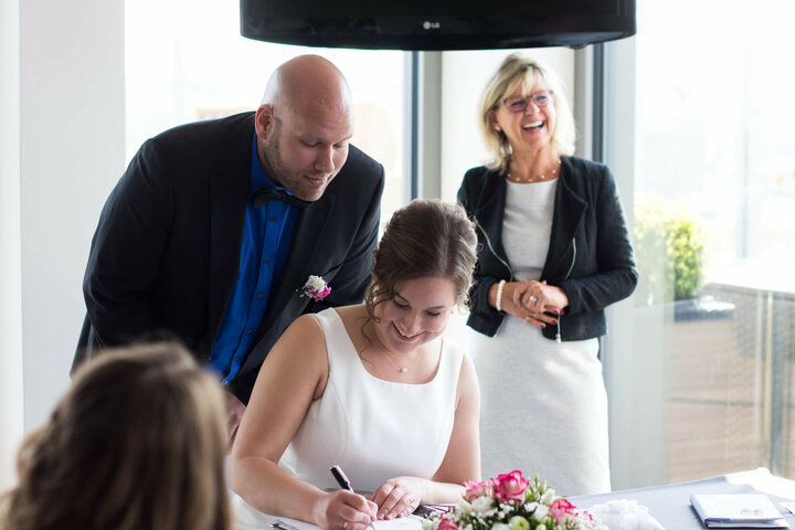Bride and groom signing documents while a smiling woman stands in the background at the ATLANTIC Hotel Kiel.