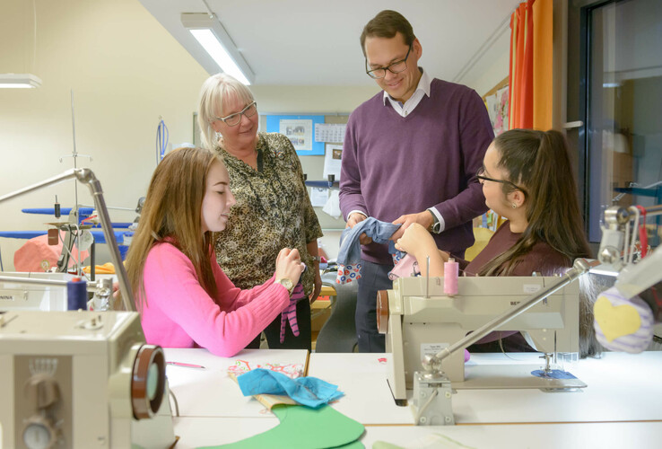 Group of people in a sewing room, two young women at sewing machines, two adults standing and watching.